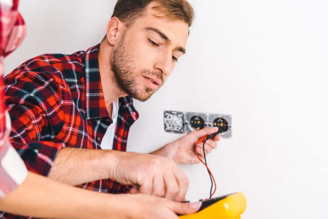 Mężczyzna naprawia gniazdko elektryczne, trzymając miernik i podłączając przewody.