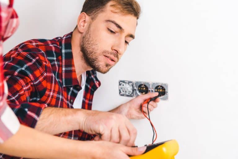 Mężczyzna naprawia gniazdko elektryczne, trzymając miernik i podłączając przewody.