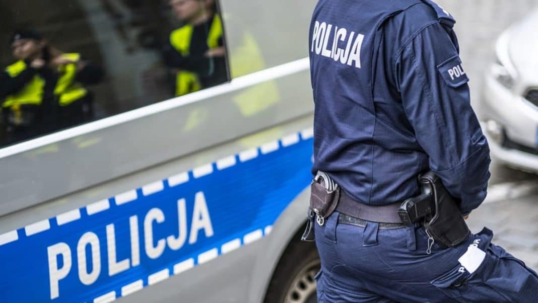 Policjant stoi obok radiowozu na tle grupy funkcjonariuszy podczas interwencji.