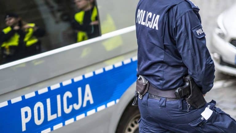 Policjant stoi obok radiowozu na tle grupy funkcjonariuszy podczas interwencji.