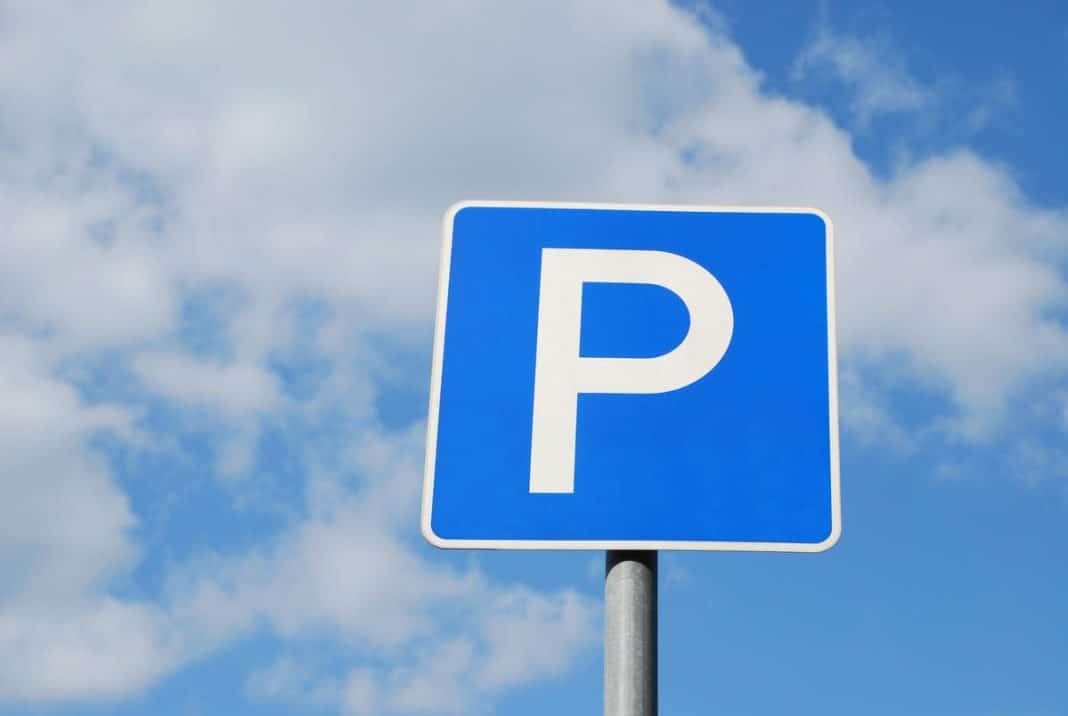 Niebieska tablica parkingowa z białym symbolem P na tle nieba z chmurami.