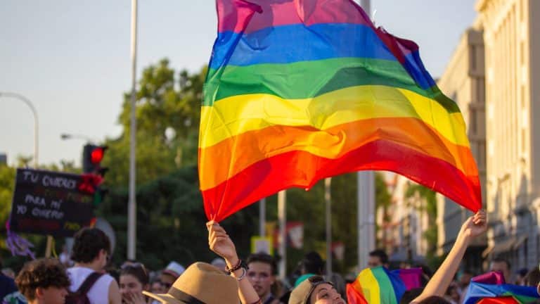 Osoba trzyma flagę w kolorach tęczy podczas wydarzenia na ulicy, w tłumie ludzi.