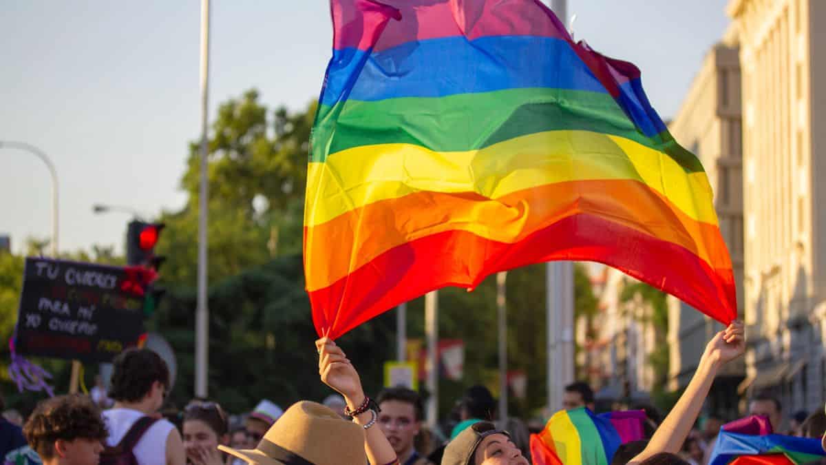 Osoba trzyma flagę w kolorach tęczy podczas wydarzenia na ulicy, w tłumie ludzi.