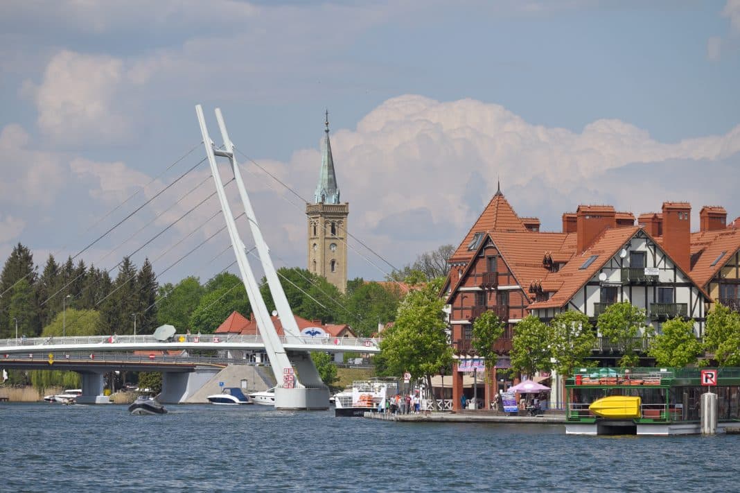 Widok na jezioro z mostem i zabytkową wieżą kościoła w tle, wśród zielonych drzew i chmur na niebie.