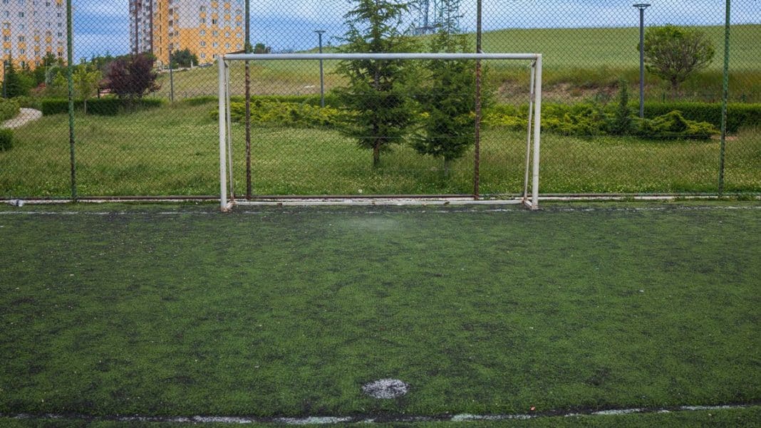 Boisko sportowe z bramką i sztuczną nawierzchnią, otoczone siatką, w tle zieleń i budynki.