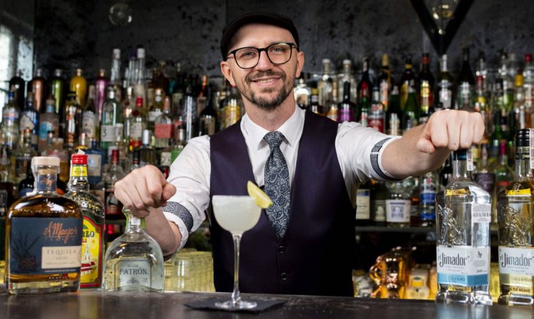 Barman w eleganckim stroju przygotowuje drinka za barem, w tle półki z różnymi butelkami alkoholu.