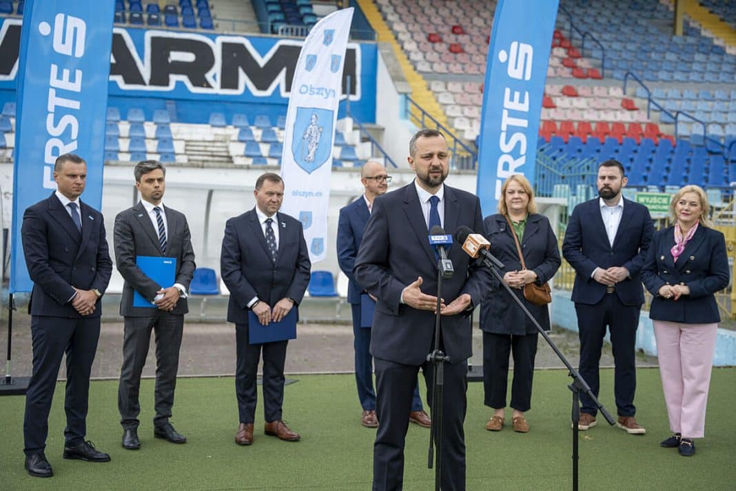 Grupa osób na stadionie podczas konferencji prasowej, z flagami i banerami w tle.