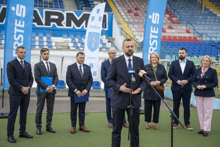 Grupa osób na stadionie podczas konferencji prasowej, z flagami i banerami w tle.