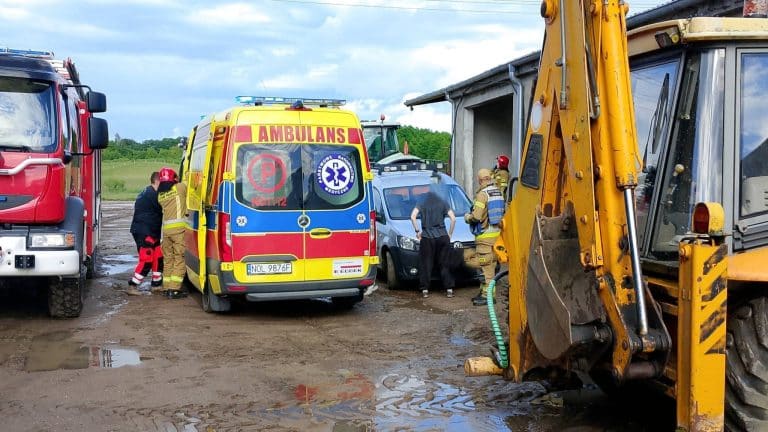 Mężczyzna przygnieciony przez łyżkę koparki podczas prac na terenie gospodarstwa, wokół służby ratunkowe i pojazdy ratunkowe.
