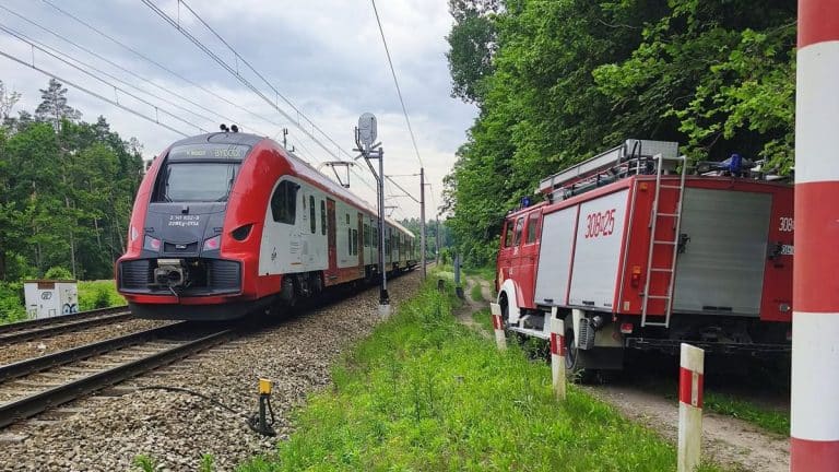 Pociąg pasażerski relacji Olsztyn-Bydgoszcz na torach w Biesalu, obok wozu strażackiego, w otoczeniu zielonych drzew.