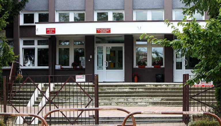 Widok na wejście do szkoły podstawowej z czerwonymi tablicami i schodami, otoczony zielenią.