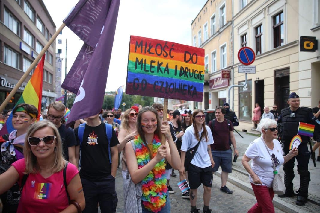 Uczestnicy marszu równości idą ulicami miasta, trzymając flagi i transparenty, w tym tęczową flagę.