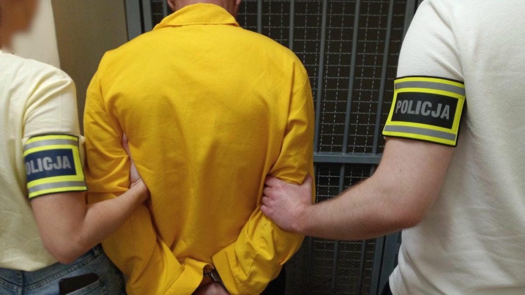 Mężczyzna w żółtej kurtce zatrzymany przez policjantów, widoczny od tyłu, w areszcie.