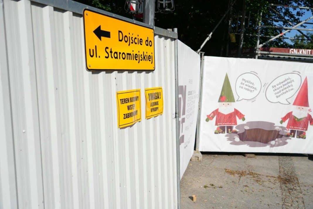 Ogrodzenie przy Wysokiej Bramie z panelami informacyjnymi i żółtymi ostrzeżeniami o głębokich wykopach.