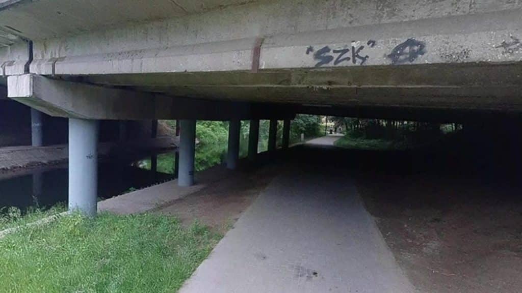 Pod mostem w parku znajduje się ścieżka rowerowa i chodnik, pod betonowym mostem widoczne graffiti.