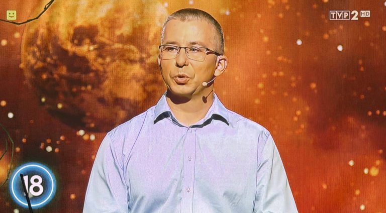 Mężczyzna w okularach i białej koszuli na tle ekranów z grafiką wybuchu atomowego.