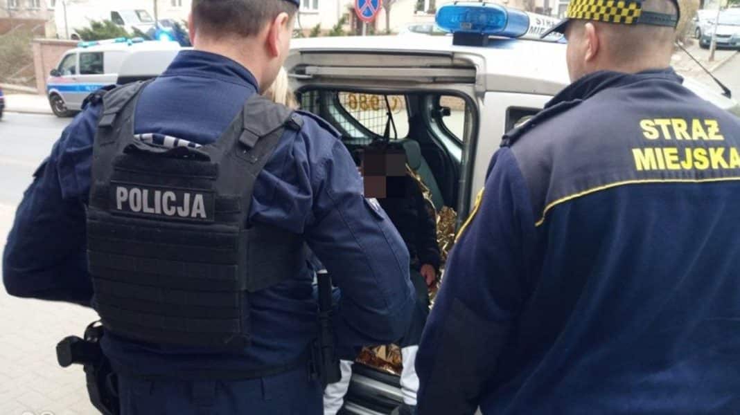 Strażnicy miejscy i policjant rozmawiają z starszym mężczyzną przy radiowozie na ulicy.