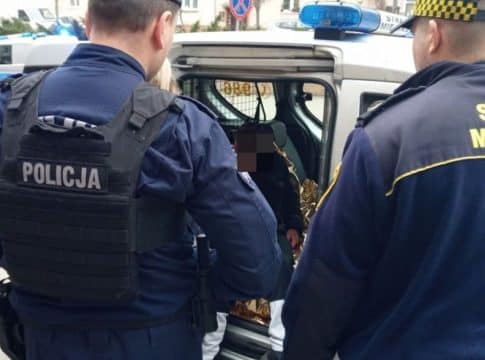 Strażnicy miejscy i policjant rozmawiają z starszym mężczyzną przy radiowozie na ulicy.