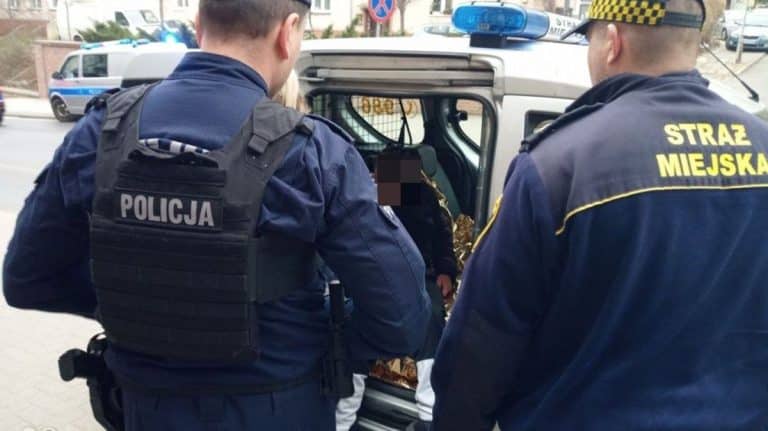Strażnicy miejscy i policjant rozmawiają z starszym mężczyzną przy radiowozie na ulicy.
