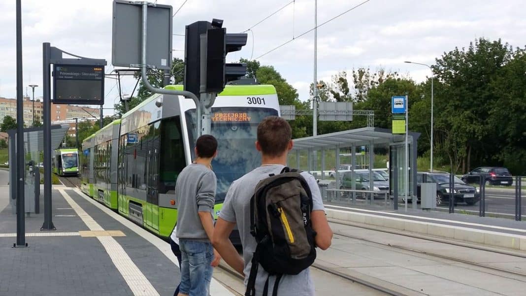 Dwie osoby czekają na tramwaj na przystanku w Olsztynie, w tle tramwaj zastępczy na torach.