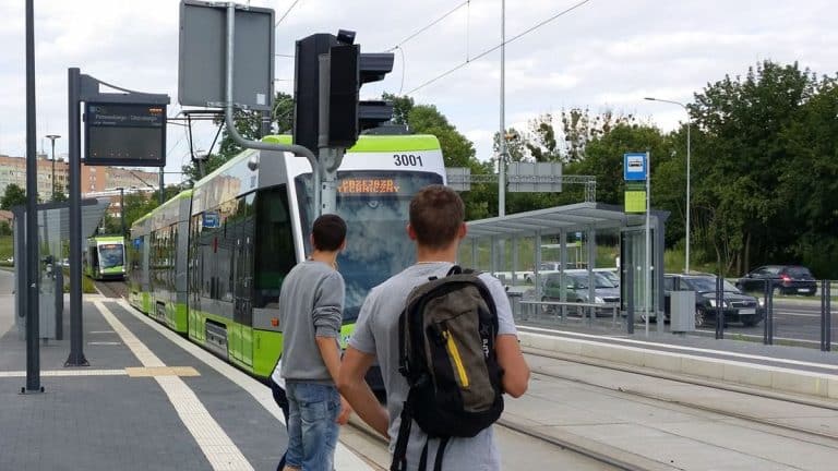 Dwie osoby czekają na tramwaj na przystanku w Olsztynie, w tle tramwaj zastępczy na torach.
