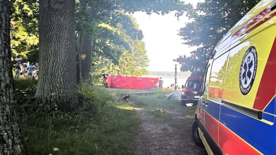 Widok na jezioro z łódką, na brzegu pojazd ratunkowy i ludzie w tle.