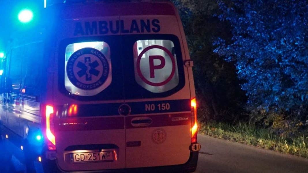Ambulans na leśnej drodze w nocy, oświetlony światłami sygnalizacyjnymi, w tle drzewa.