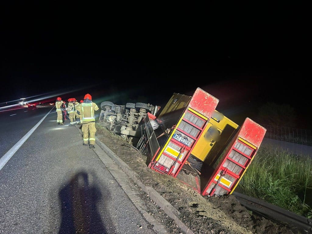 Ciężarówka leży na boku przy drodze, wokół niej strażacy podczas akcji ratunkowej, nocne warunki.