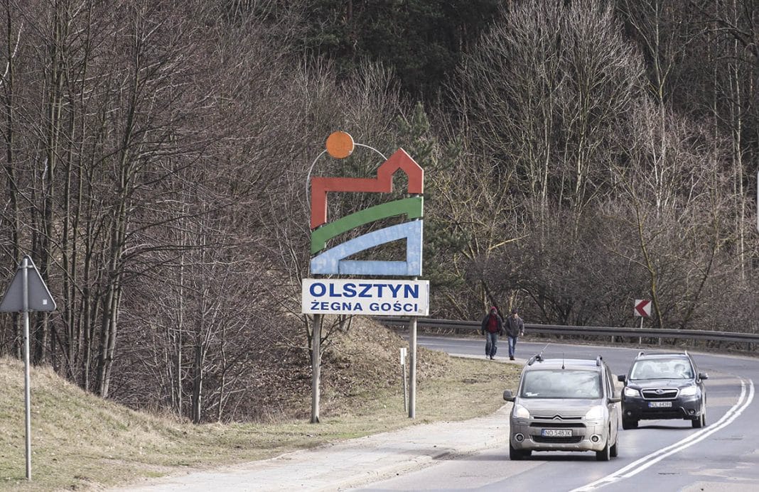 Tablica powitalna przy wjeździe do Olsztyna z napisem 'Olsztyn żegna gości', otoczona lasem i samochodami na drodze.