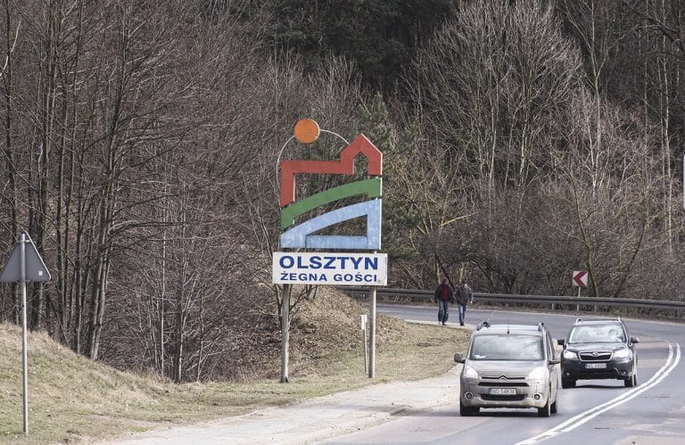 Tablica powitalna przy wjeździe do Olsztyna z napisem 'Olsztyn żegna gości', otoczona lasem i samochodami na drodze.