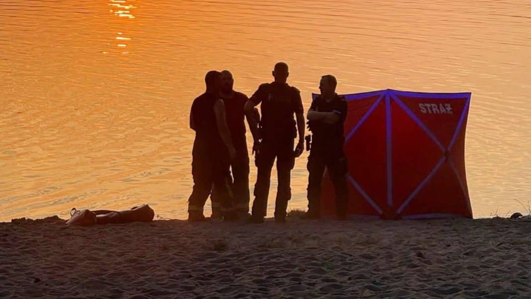 Grupa ratowników i strażaków na plaży nad jeziorem o zachodzie słońca, z namiotem ratowniczym.