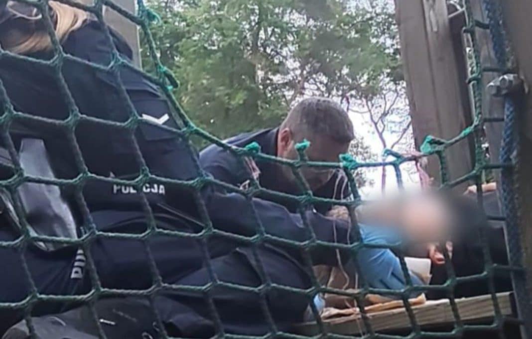 Policjanci udzielają pomocy dziecku z raną głowy w parku, widoczny ogrodzenie i drzewa w tle.