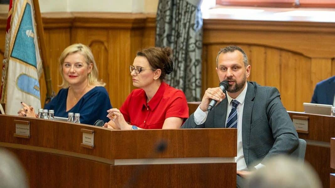 Trzech urzędników siedzi za biurkiem podczas sesji Rady Miasta Olsztyna, jeden z nich mówi do mikrofonu.