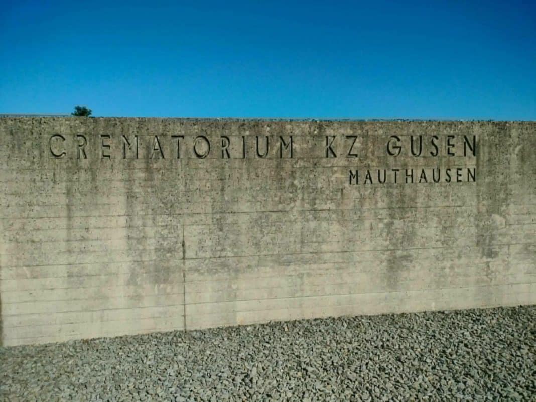 Betonowa ściana z napisem 'Cmentarz KZ Gusen' na tle nieba.
