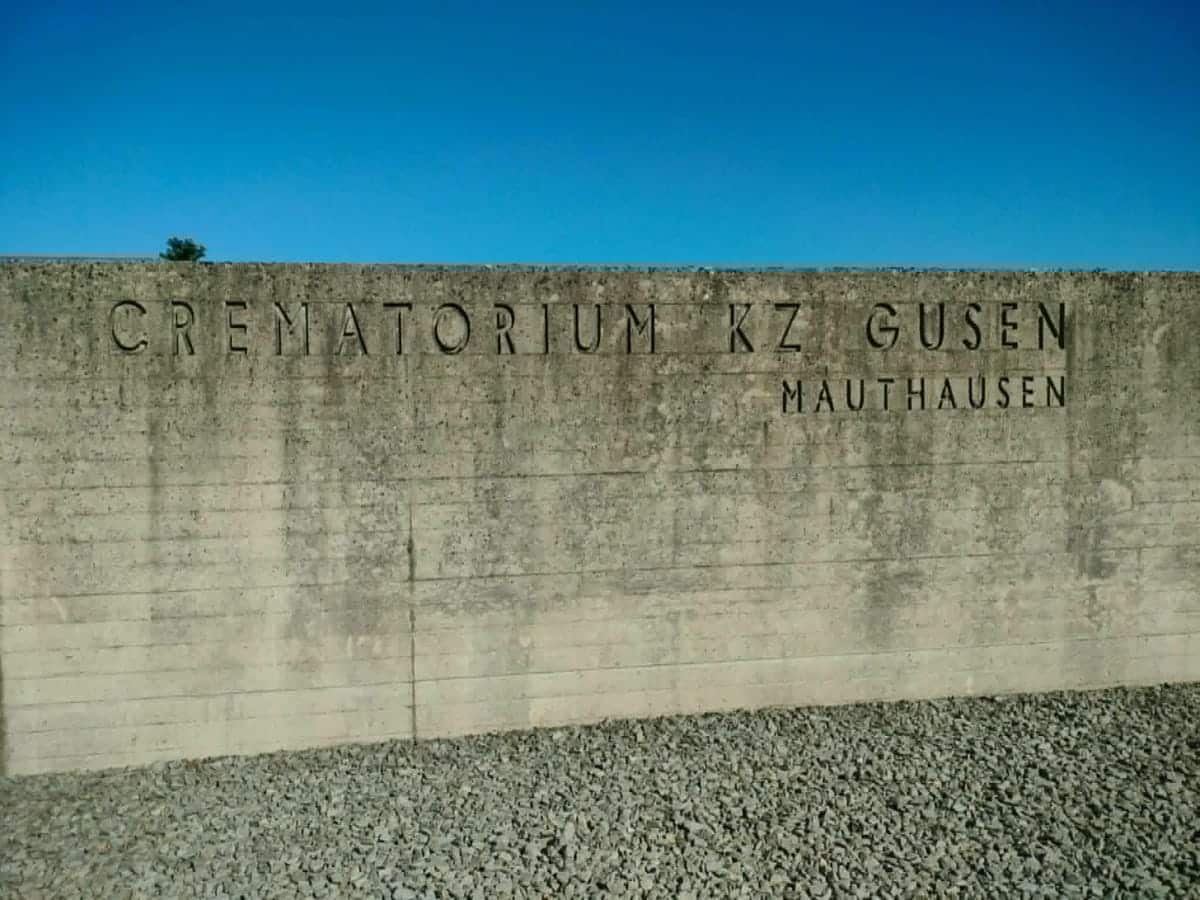 Betonowa ściana z napisem 'Cmentarz KZ Gusen' na tle nieba.