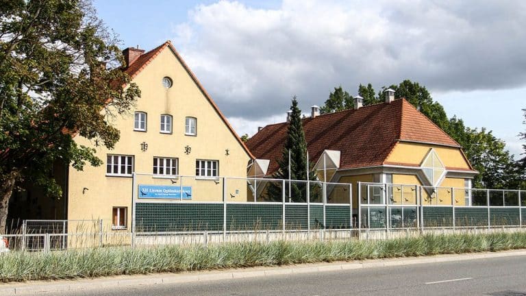 Szkoła średnia w Olsztynie z budynkami i ogrodzeniem przy ulicy, pod chmurami.