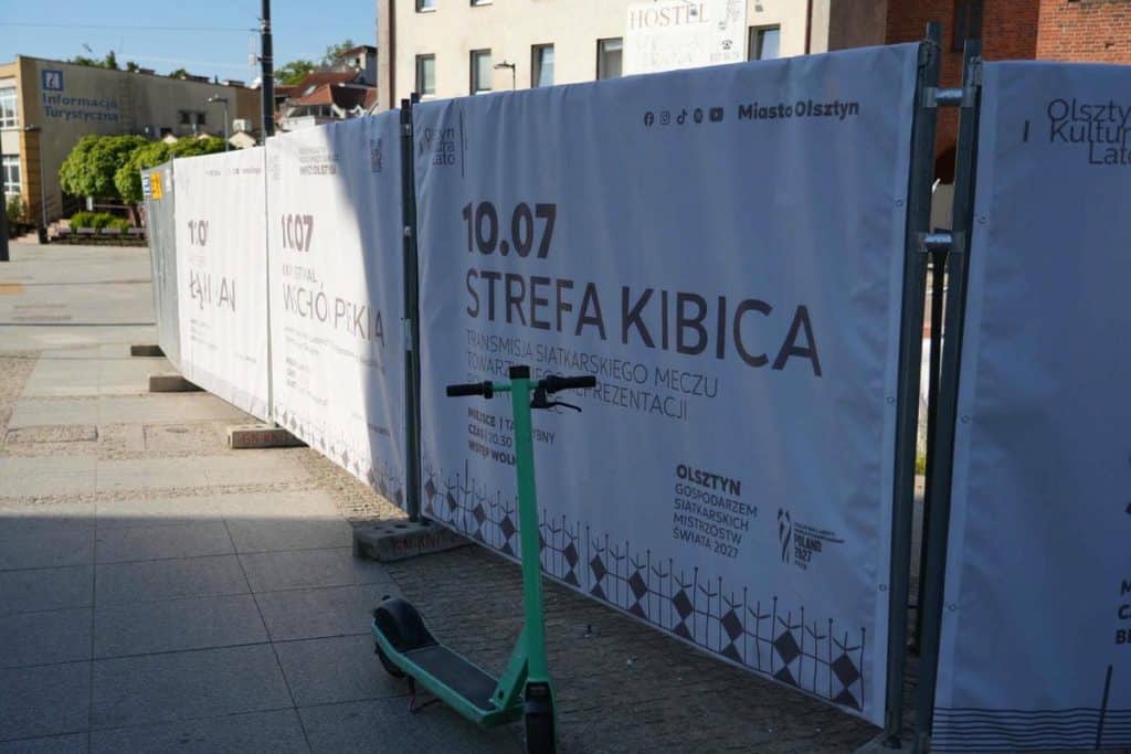 Płot na Starym Mieście w Olsztynie z panelami informacyjnymi, w tle budynki i niebo.