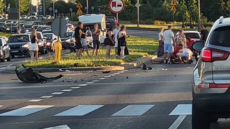 Grupa ludzi czeka na miejscu wypadku na skrzyżowaniu, widoczne rozbicie motocykla i samochodu.