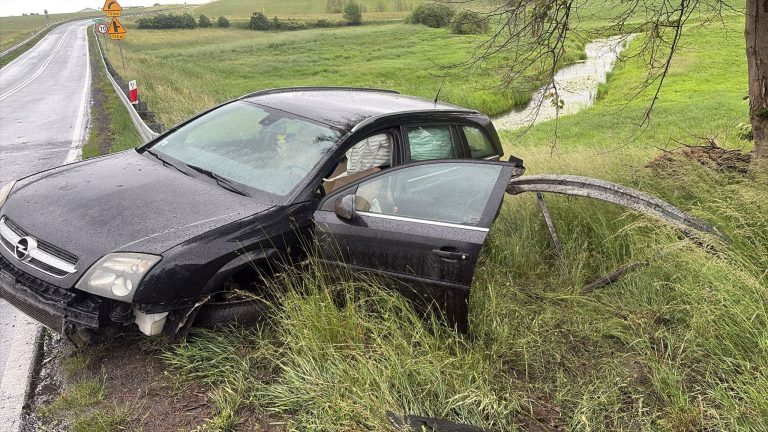 Rozbity czarny samochód Opel na poboczu drogi, w pobliżu rowu, po kolizji na mokrej nawierzchni.