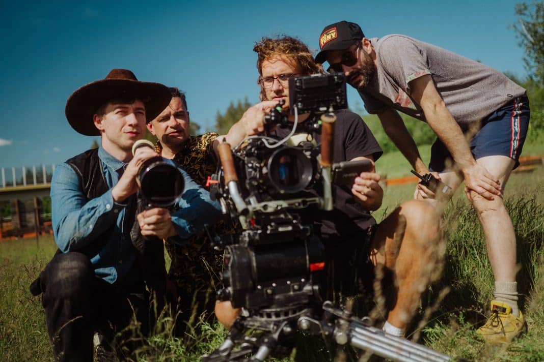 Grupa ludzi na planie filmowym w miasteczku westernowym, obsługa kamery i aktorzy w kostiumach.