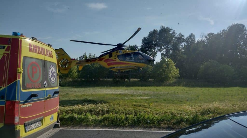 Helikopter ratunkowy ląduje na polu po wypadku na DK16, obok ambulansu, w tle drzewa i niebo.