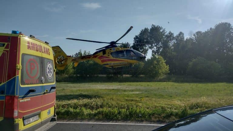 Helikopter ratunkowy ląduje na polu po wypadku na DK16, obok ambulansu, w tle drzewa i niebo.