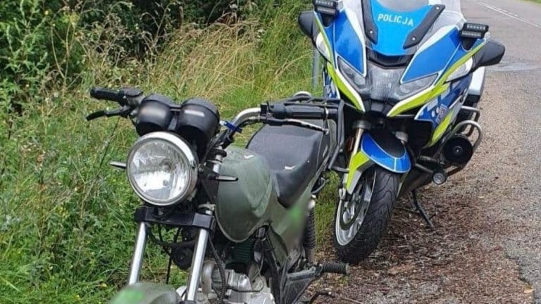 Motocykl policyjny i motorower na poboczu drogi, otoczone zaroślami.