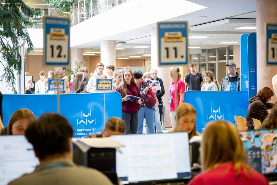 Grupa kandydatów czeka przy stanowiskach rekrutacyjnych na uczelni, w tle widoczne tablice z numerami punktów rekrutacji.