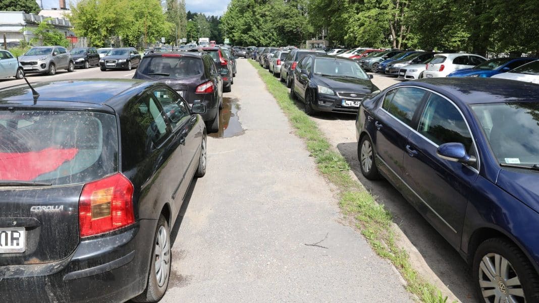 Zapełniony parking samochodami przy ulicy, w tle drzewa i budynki.