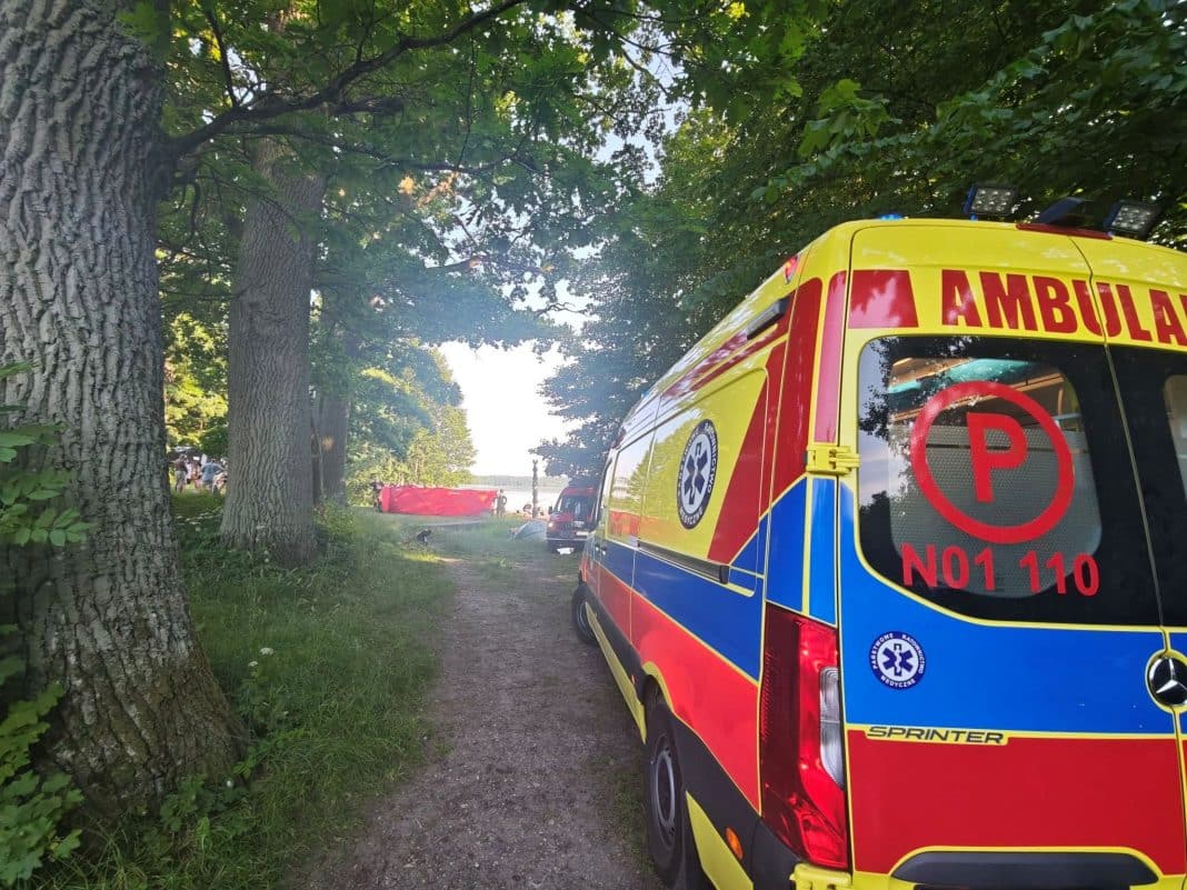 Na zdjęciu widoczny jest ambulans na tle drzew i jeziora, w tle grupa ludzi przy brzegu.