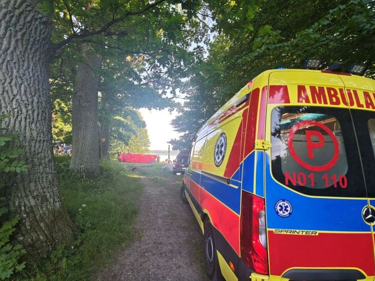 Na zdjęciu widoczny jest ambulans na tle drzew i jeziora, w tle grupa ludzi przy brzegu.