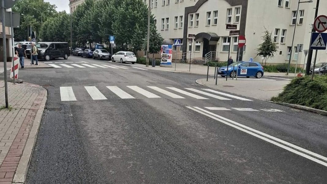 Widok na przejście dla pieszych na ulicy w Olsztynie, z oznaczeniem poziomym i sygnalizacją świetlną.