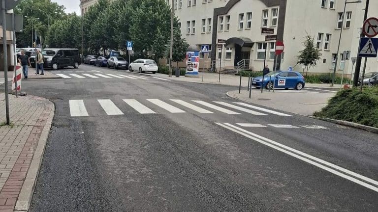 Widok na przejście dla pieszych na ulicy w Olsztynie, z oznaczeniem poziomym i sygnalizacją świetlną.