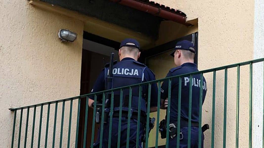 Dwóch policjantów stoi przy wejściu do domu, rozmawiając, z widocznymi oznaczeniami 'Policja' na mundurach.